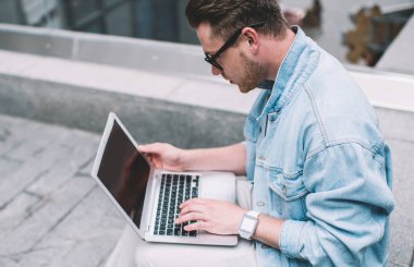 Gözlüklü ve kot ceketli havalı beyaz adam sokak çitlerinde oturuyor ve taşınabilir laptop üzerinde çalışıyor.