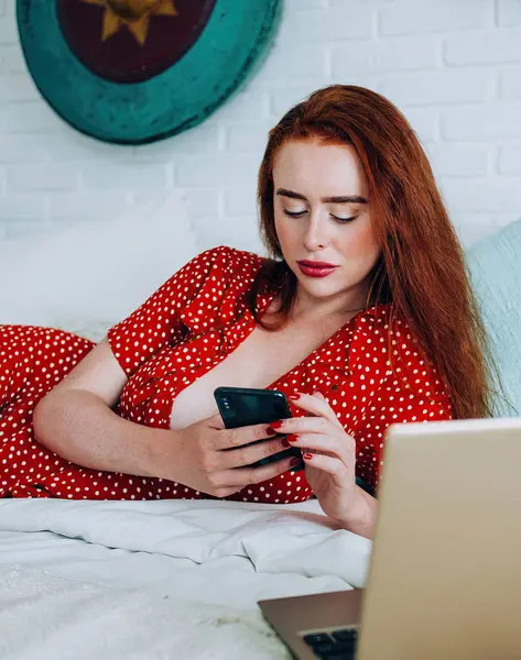 Focused woman with red hair in red polka dot dress lying on bed near laptop and texting on smartphone at home