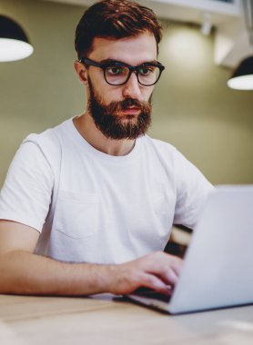 Gözlüklü ciddi bir erkek blogcu dizüstü bilgisayarda yayınlanması için makale yazıyor, yetenekli hipster adam internetteki internet dersini birlikte çalışma odasında izlemeye odaklanmış.
