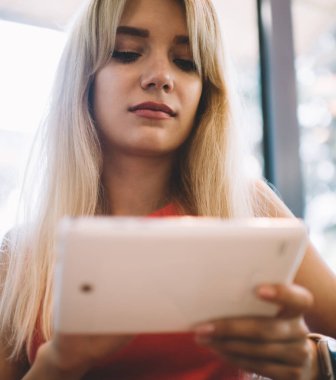 Düşük açılı, sarı saçlı, tablet kullanan, pencereye yaslanmış, rahat, modern bir kafede dinlenen bir kadın.