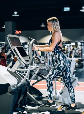 Spor salonunda spor yapan, kamuflaj eşofmanlı genç ve sarışın bir kadının spor salonunda spor yaparken sağlıklı yaşam tarzının keyfini çıkarması.