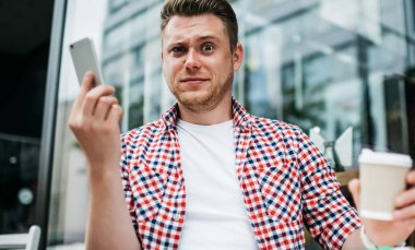 Bin yıllık asık suratlı adam elinde telefon ve kahve fincanı tutarken sokakta oturmuş şaşkınlık ifadesiyle kameraya bakıyordu.