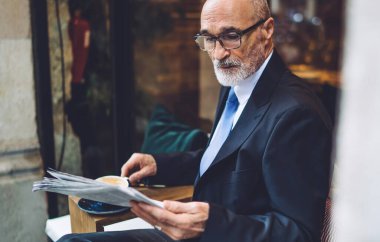 Sakallı, kahve bardaklı, takım elbiseli, beyaz duvarın yanında oturan ve gazete okuyan yaşlı adamın yan görüntüsü.