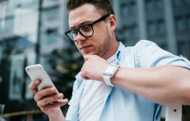 Akıllı saat ve gözlüklü yakışıklı milenyum erkeğinin sokak terasında otururken konsantrasyonlu akıllı telefon kullanmasından.