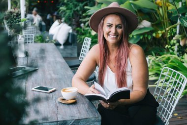 Kafe terasında kitap okurken elinde kahve fincanıyla oturmuş kameraya bakan şapkalı çekici bir kadın.