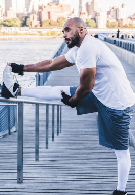 Kendine güvenen Afrikalı Amerikalı sporcunun sabah saatlerinde demir çitlerin üzerinde bacaklarını esneterek arka plandaki deniz manzarasına bakması.