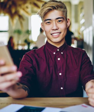 4G interneti kullanarak akıllı telefon cihazının profilini güncellerken gülümseyen Çinli pozitif hipsterin portresi. Masaüstünde oturan yakışıklı Asyalı öğrenci cep telefonu tutuyor.