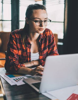 Kafeteryadaki masada oturmuş kağıt çizelgelerle çalışırken taşınabilir dizüstü bilgisayarları karıştıran gözlüklü esmer bir genç.