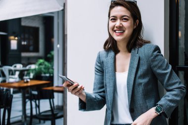 Genç, çekici, güzel, neşeli, resmi giyinmiş, akıllı telefon kullanan, duvara yaslanmış, caddede kameraya bakan bir kadın.