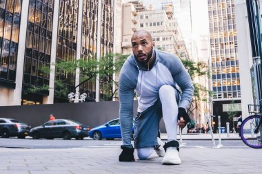 Spor kıyafetleri ve eldiven giymiş yakışıklı Afro-Amerikan koşucu yol kenarında diz çöküp, antrenmanda başını çevirirken kulaklık takarak müzik dinliyor.