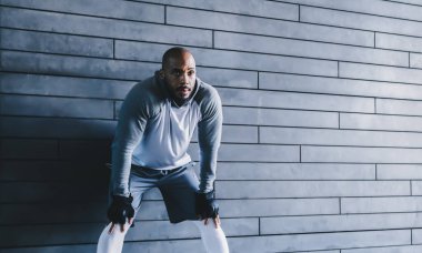 Gri spor giyimli, kel, yorgun ve sakallı bir adam. Duvara yaslanmış siyah eldivenleri var. Elleri dizlerinin üstünde ve dinlenirken başka yere bakıyor.