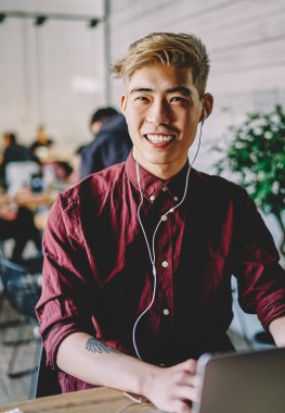 Modern laptop cihazına bağlı kulaklıkla müzik dinleyen olumlu Japon genç adam kablosuz internet kullanıyor. Yaratıcı Çinli hippi adam web sitesinden ses şarkıları dinlemekten hoşlanıyor.