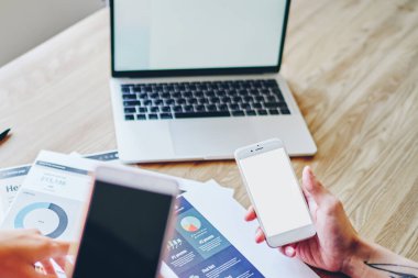 İnternet siteniz için fotokopi alanı olan modern akıllı telefonları tutan genç adam ve kadının kırpılmış görüntüsü veya ahşap masaüstünde dizüstü bilgisayarlı ve diyagramlı kağıt çizimleriyle oturan içerik