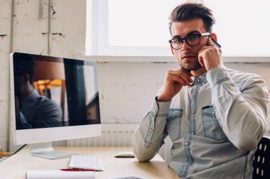 Ofiste modern bilgisayarla masaüstünde otururken akıllı telefon cihazıyla konuşan gözlüklü ciddi bir gencin portresi. Telefon cihazıyla konuşan sıradan giyinmiş bir erkek öğretmen.