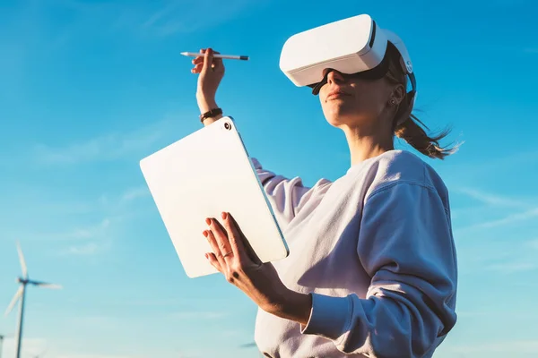Low angle of woman immersed in the VR game and looking around in white goggles while creating digital virtual reality space with touchpad program and stylus
