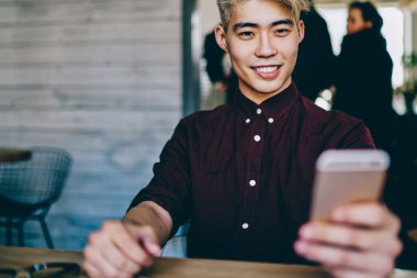 Neşeli Çinli yenilikçi adam sosyal ağlarda akıllı telefondan komik videolar izliyor modern mekanda oturuyor. Japon blogcu kendi web sitesinden 4G internet kullanarak yayın indiriyor.