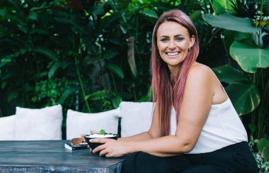 Tarz sahibi pembe saçlı, neşeli yetişkin bir kadın. Dostça gülümseyen ve kameraya bakan. Tropikal bahçede kahvaltıda sağlıklı yulaf lapasının tadını çıkaran.