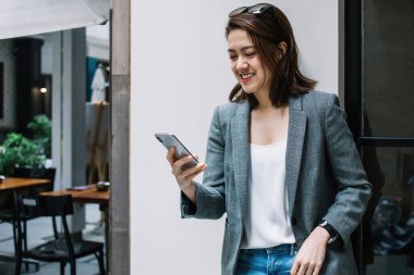 Güzel, güzel, mutlu, resmi giyinmiş, akıllı telefon kullanan, şehrin sokaklarında duvara yaslanmış, gülen bir kadın.