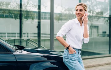 Şen şakrak sarışın bayan sürücü akıllı telefondan konuşuyor ve gülümsüyor. Siyah arabanın yanında duruyor ve şapkaya yaslanıyor.