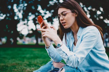 Çevreci genç bir kadın yeşil çimlerde oturuyor ve internet sitesinde haber okuyor modern akıllı telefon cihazında. Sosyal ağlarda sosyal medyada sohbet eden hippi kız şehrin ulusal parkında cep telefonuyla konuşuyor.