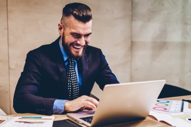 Resmi giyinmiş pozitif sakallı CEO modern bilgisayarda komik webinar izlerken kablosuz internet kullanıyor. Ofisteki dizüstü bilgisayardaki video çağrısında gülen takım elbiseli başarılı bir girişimci.