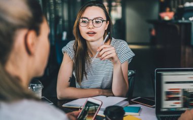 Gözlüklü hoş bir kadın masada iş arkadaşlarıyla oturuyor, not defterleri ve cihazları var ve kafede buluşurken sohbet ediyorlar.