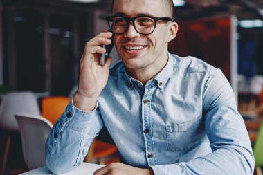 Modern çalışma ortamında masada oturan gözlüklü, akıllı telefonlu bir arkadaşla iletişim kuran pozitif hipster. Kot gömlek giymiş neşeli genç adam cep telefonundan ararken gülüyor.