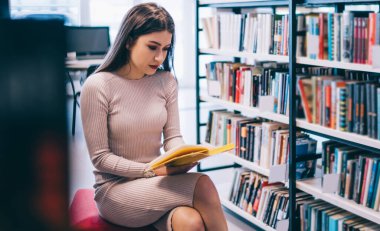 Modern üniversite kütüphanesinde kitaplığın yanında oturup ders çalışırken şık giyinmiş, ilginç kitaplar okuyan güzel bir kız öğrenci.