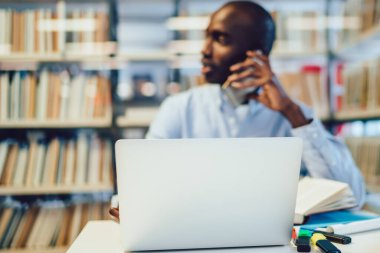 Çeşitli kırtasiye ve modern dizüstü bilgisayar kütüphane masasının yanına yerleştirilmiş. Bulanık Afrikalı Amerikalı bir adam akıllı telefondan kitaplığın yanında konuşuyor.