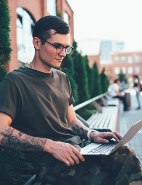Gözlüklü ve sıradan kıyafetleri olan genç bir adamın ara sokakta bankta otururken dizüstü bilgisayarda yazışının yan görüntüsü.