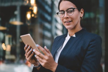 İçerik siyah saçlı, şık giyinmiş, gözlüklü bir kadın şehir sokaklarında dijital tablet hakkında bilgi ararken kameraya bakıyor.
