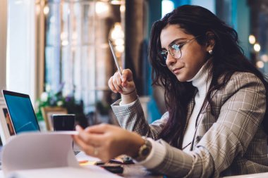 Kapalı ceketli, koyu uzun saçlı konsantre bir kadın kahve dükkanında dizüstü bilgisayarla masada oturan not defterinin sayfalarını çeviriyor.
