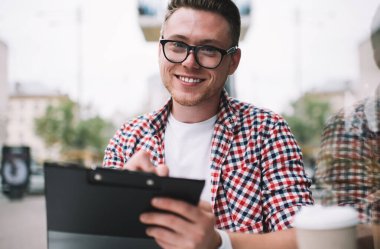 Ekose gömlekli ve gözlüklü mutlu bir beyaz adam, kafeterya masasında kameraya gülümserken panoya yazı yazıyor.