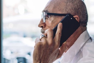 Beyaz gömlekli, gözlüklü, kol saati takmış akıllı telefondan konuşan ve pencereden arka plandaki güneş ışığına bakan sakallı yaşlı adamın yan görüntüsü.