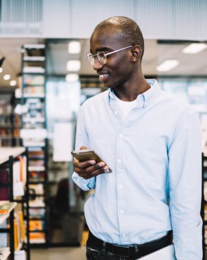 Akıllı, yakışıklı, gözlüklü, gülümseyen cep telefonlu, kitaplıklarda yürürken okumak için kitap seçen Afro-Amerikan bir adam.