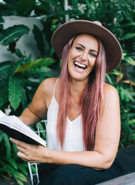 Çekici, şapkalı, boyalı saçlı, kitap okuyan, tropik çalıların yanında oturan ve kameraya bakan bir kadın.