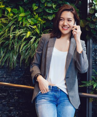 Mutlu, genç, gülümseyen, şık kıyafetler içinde, gri ceketli, mavi kotlu, telefonda konuşan güzel Asyalı kadın.