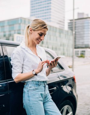 Beyaz bluzlu neşeli sarışın kadın ve kotlar cep telefonunu karıştırıyor ve bulanık şehir arka planında siyah arabaya yaslanıyor.