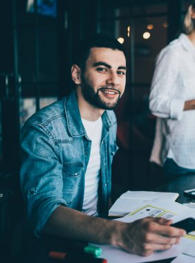 Kot gömlekli sıradan bir adam elinde kağıtlar ve not defterleriyle masada oturuyor ve arka planda bulanık kadın iş arkadaşlarıyla kameraya gülümsüyor.
