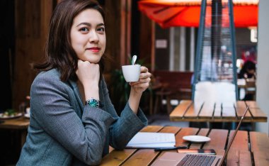 Genç Asyalı bir kadın elinde bir fincan kahveyle kameraya bakarken elinde dizüstü bilgisayarla ahşap masada oturuyor.