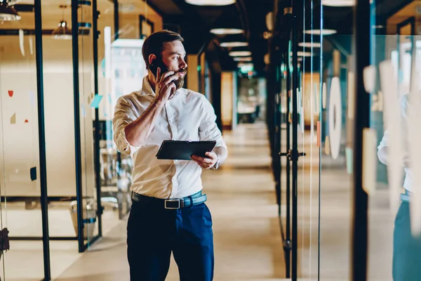 Caucasian bearded businessman in formal wear looking away while discussing plan of new project during phone call on smartphone device.Entrepreneur standing in office corridor and talking on cellphone