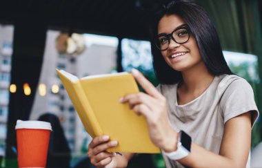 Elinde ilginç bir kitap tutan neşeli beyaz kadın portresi hobi için boş vakti olmasından memnun, gözlüklü yeniyetme kız kafede kameraya bakarak en çok satanlardan birinin keyfini çıkarıyor.