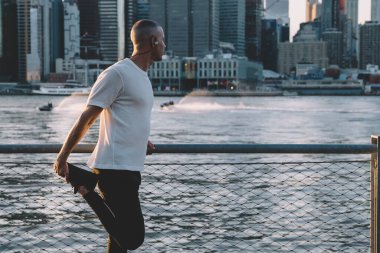 Spor giyim ve kulaklıklı genç bir erkeğin yan görüntüsü. Şehir arka planında gökdelenlerle şehir manzarasına bakarken bacağını uzatan kulaklıklar.