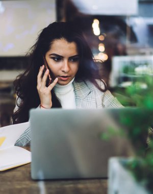 Uzun siyah saçlı, rahat giyimli, cep telefonuyla konuşan ve dizüstü bilgisayarla masada oturan zeki bir kadın.
