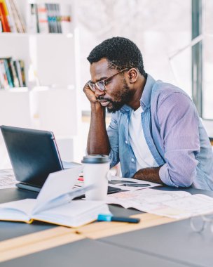 Gözlüklü, tişörtlü, dizüstü bilgisayarlı bir Afro-Amerikan adam gözlüklü, not defterleriyle dolu bir masada oturuyor.