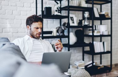 Ciddi bir beyaz erkek kredi kartı bilgilerini kontrol ediyor bilgisayarla işlem yapıyor ve evdeki kablosuz bağlantı kuruyor, netbook 'u elinde tutan konsantre bir hipster kanepeye oturmuş online alışveriş yapıyor.