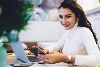 Kafeteryada oturmuş kameraya bakarken dizüstü bilgisayar ve cep telefonu kullanarak projede çalışan neşeli, genç esmer kadın.