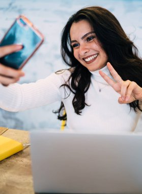 Genç, neşeli bir kadın cep telefonuyla selfie çekiyor ve kafeteryadaki derslere hazırlanırken parmaklarıyla barış işareti gösteriyor.