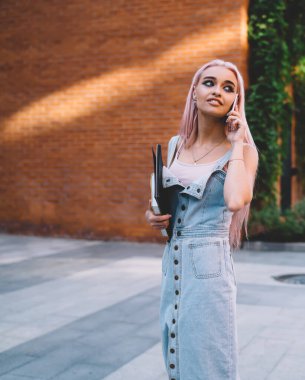 Gülümseyen ve arkasını kollayan defterleri olan neşeli genç bir kadın kaldırımda dikilip telefonlarına cevap veriyor.