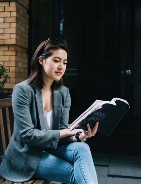 Genç, çekici, odaklanmış ve resmi kıyafet giymiş Asyalı bir kadın. Şehir caddesinde bankta otururken ilginç bir şekilde kitap okuyor.